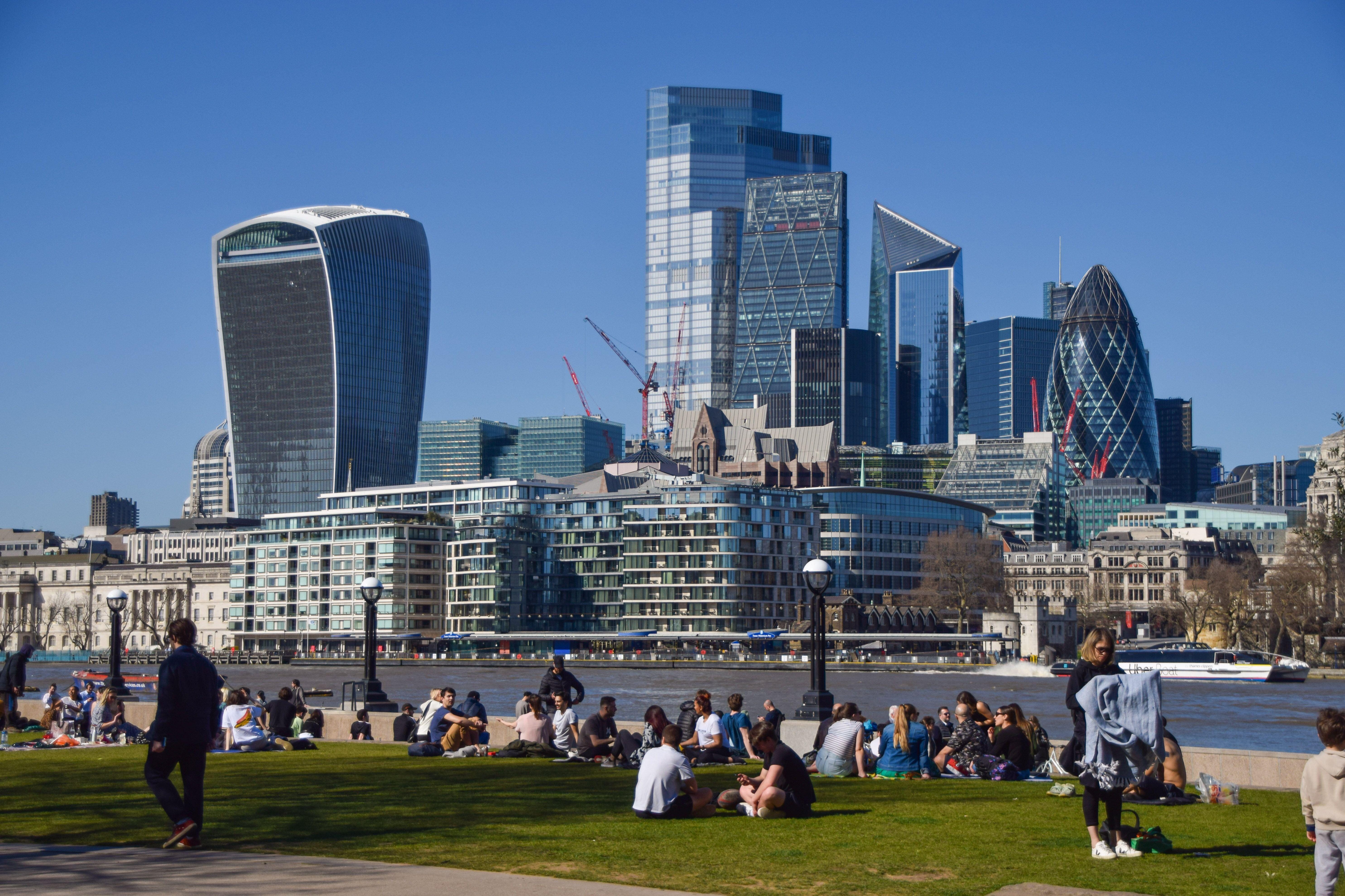 Individuals enjoying sunny weather at Potters Fields Park with Tower Bridge and the City of London skyline in the background.